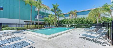 A pool area with lounge chairs and a table, surrounded by a fence and palm trees.