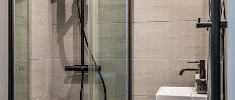 Ducha con cerramiento de vidrio, cabezal de ducha de mano, grifo empotrado y lavamanos blanco con toallero.