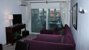 Un salon avec un canapé violet, un téléviseur posé sur un meuble en bois et une porte vitrée donnant sur un balcon avec vue sur la ville.