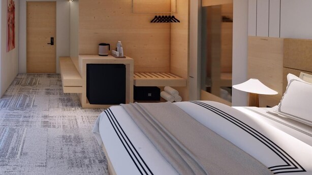 Ein modernes Hotelzimmer mit einem großen Bett, einer kleinen Sitzecke, einer Holzwand mit Ablagefläche und einer Glastür zum Badezimmer.
