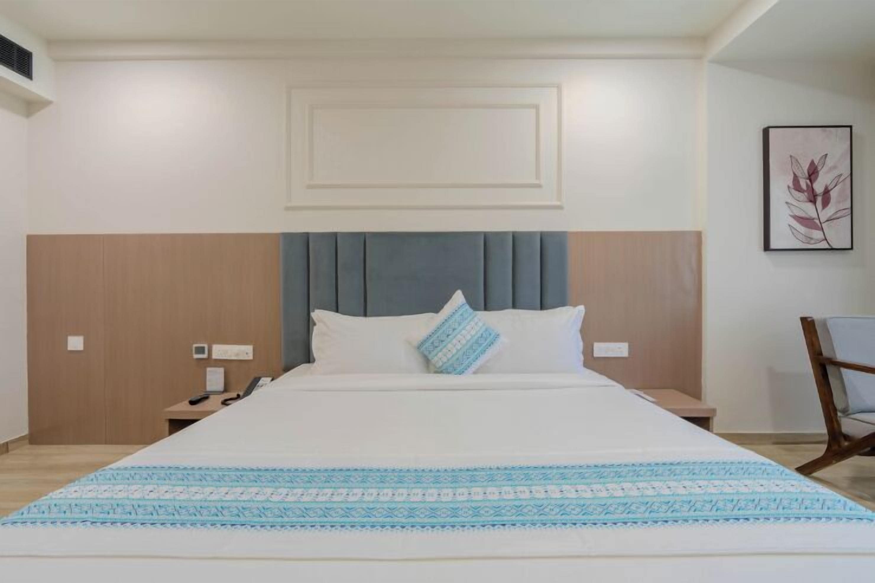 Habitación de hotel con una cama grande, cabecero, mesita de noche, una silla y una obra de arte colgada en la pared.