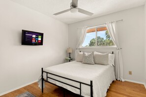 Un dormitorio con cama, ventilador de techo, ventana con cortinas y televisor montado mostrando servicios de streaming.