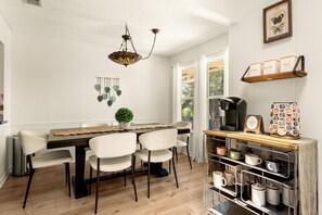 Ein Essbereich mit Holztisch, weißen Stühlen, einer Hängeleuchte und einer Kaffeestation mit Tassen und einer Kaffeemaschine.