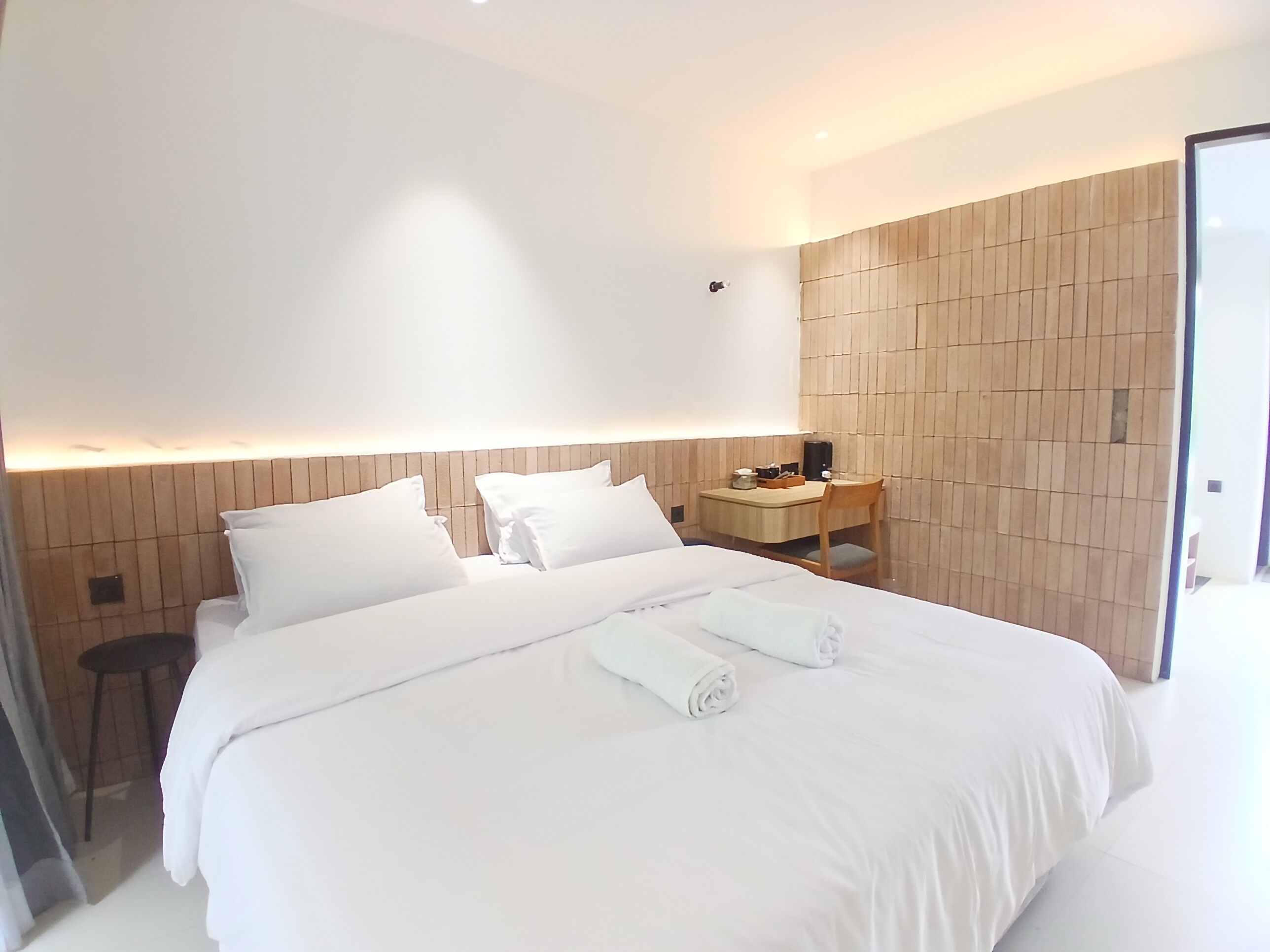 Une chambre d’hôtel moderne avec un grand lit, une tête de lit en bois, un petit bureau avec une chaise et une table de chevet avec une lampe.