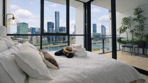 A modern bedroom with a city view, a bed with white linens, a hat, and a small table with a plant. - Million Dollar Views Luxury Apartment with 3pm Late Checkout (South Brisbane)