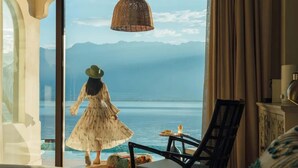 A room with a large window overlooking a body of water and mountains. There is a chair and a table with a candle and a plate of food. - Dali Pure-Soul Seaview Resort Hotel (Dali)