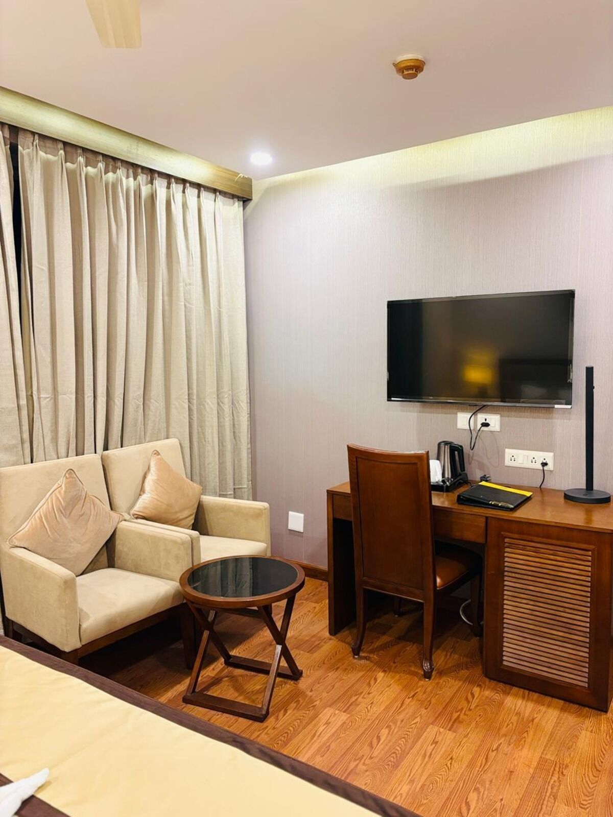 A hotel room with a sofa, a small round table, a desk with a chair, a television mounted on the wall, and a ceiling fan.