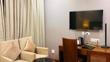 A hotel room with a sofa, a small round table, a desk with a chair, a television mounted on the wall, and a ceiling fan.