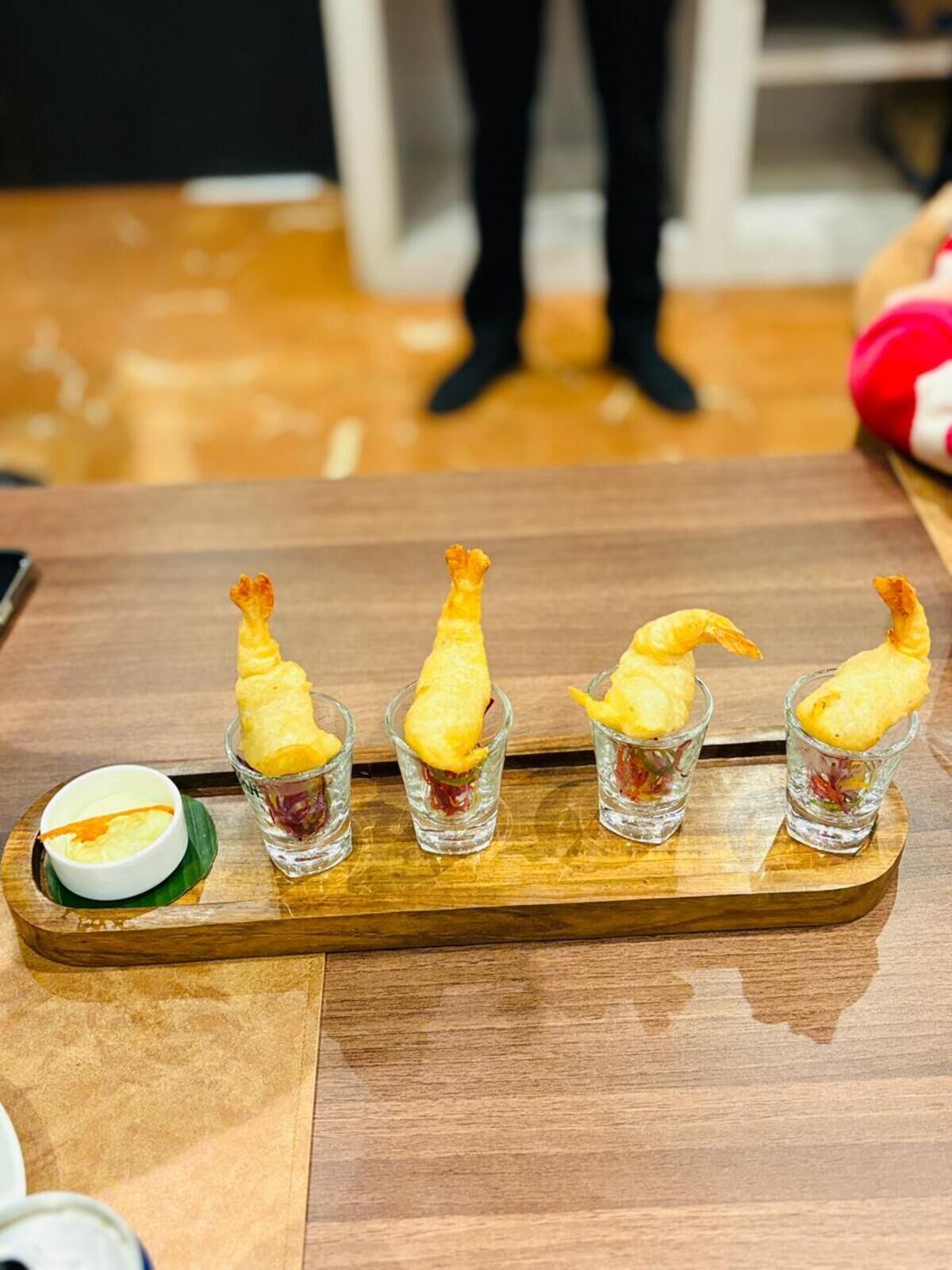 A wooden table with four shot glasses containing a golden-brown item, possibly fried food, and a small bowl of sauce on a green leaf.