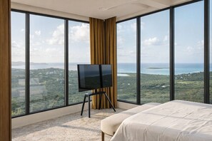 Ein Schlafzimmer mit einem großen Bett, einem Fernseher auf einem Schrank und Blick auf eine Landschaft durch große Fenster.
