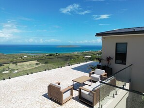 Um terraço com mobiliário para área externa, guarda-corpo de vidro e vista para o mar.