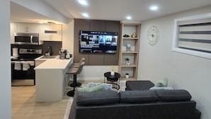 Een moderne woonkamer met een grote flatscreen televisie aan de muur, een donkergrijze bank en een keukenhoek met een magnetron en oven.