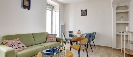 A modern living room with a green sofa, a coffee table with a bottle and glasses, and a dining area with a round table and colorful chairs.