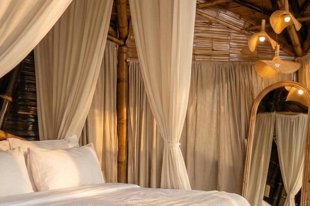 Una cama con sábanas blancas, un espejo grande y cortinas con drapeados en una habitación con vigas de bambú.