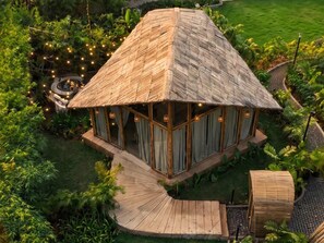 A thatched-roof structure with large windows, surrounded by lush greenery and a wooden deck.