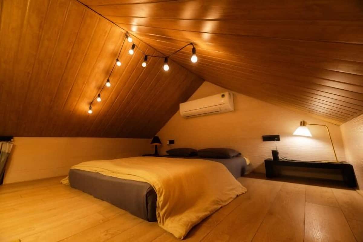 Un dormitorio acogedor en el ático, con techo inclinado, cama, lámparas de noche y un equipo de aire acondicionado.