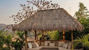 A thatched-roof outdoor seating area with cushions, surrounded by lush greenery and a view of the landscape. - Whispering Woods (Bhiwandi)