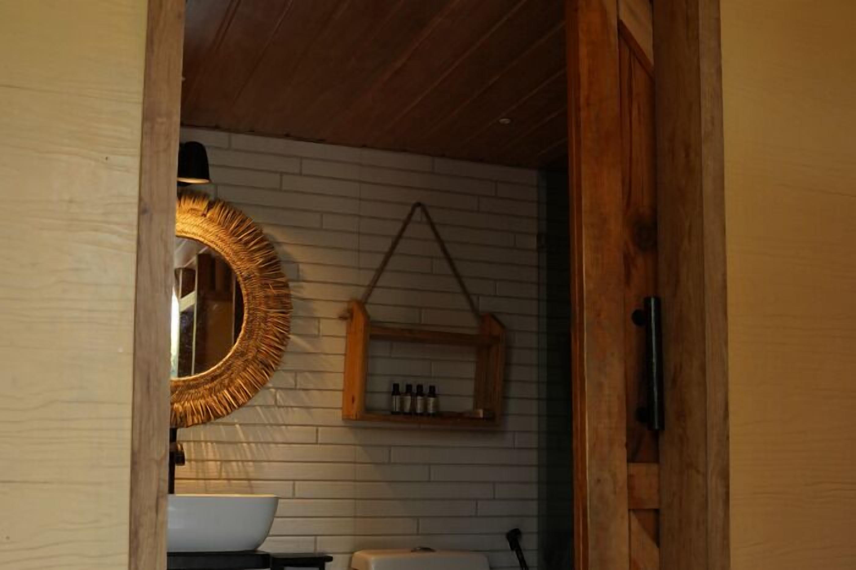 Un baño con pared de ladrillo blanco, una puerta de madera, un espejo redondo, un estante con botellas y un lavamanos blanco.
