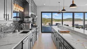 A modern kitchen with a large island, stainless steel appliances, and a view of a beach through large windows. - Lakefront resort home. Sleeps 26+! Boat dock, kayaks, pool, hot tub & more! (Graham)
