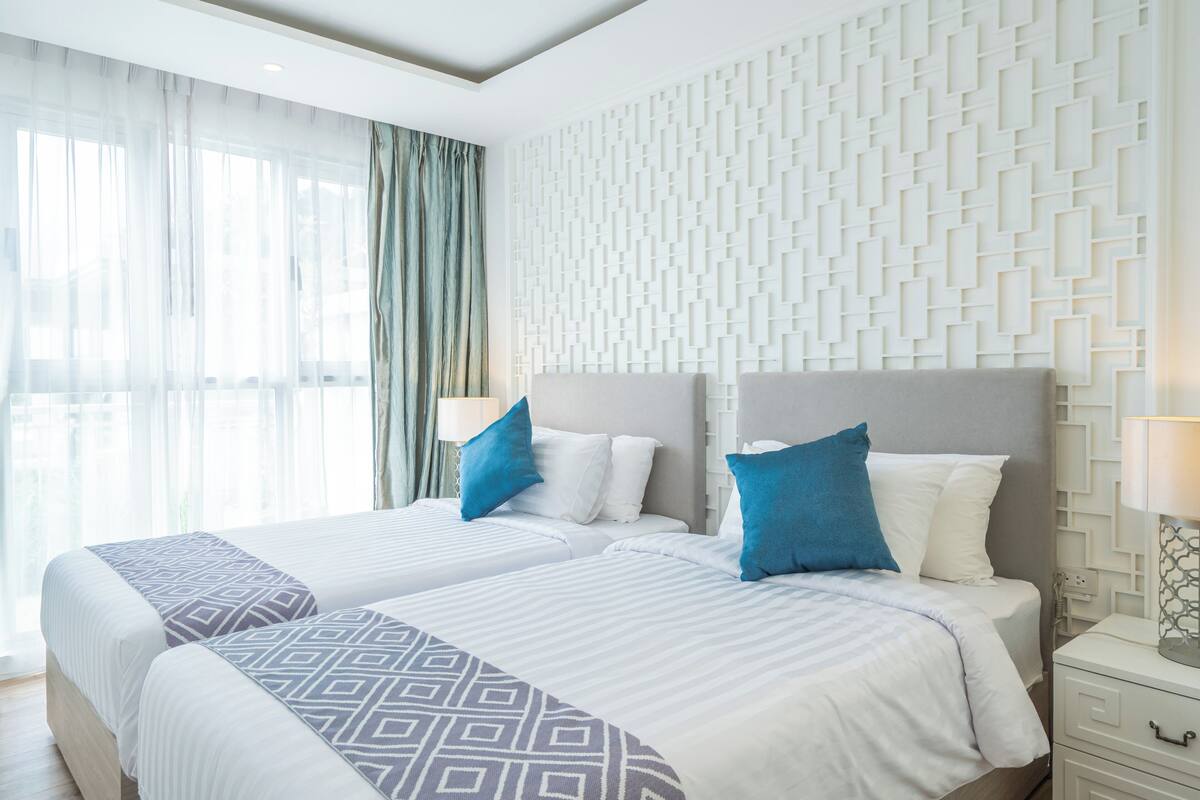 A hotel room with two beds, a patterned headboard, a geometric wall design, and a window with sheer curtains.