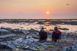 Two people sitting on chairs facing a rocky beach at sunset.