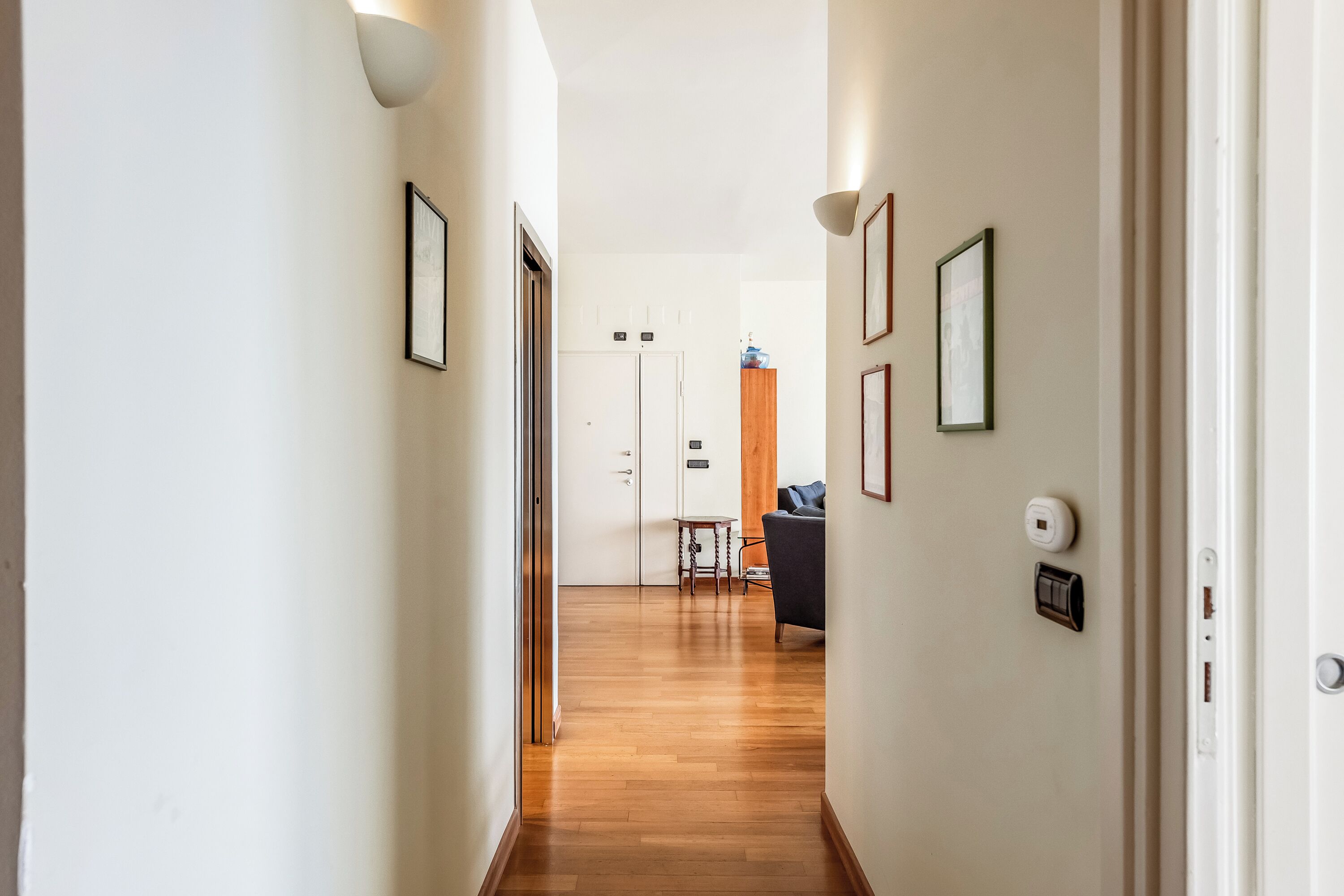 A hallway with wooden flooring, white walls, and framed pictures. There is a door at the end leading to a room with a sofa and a table.