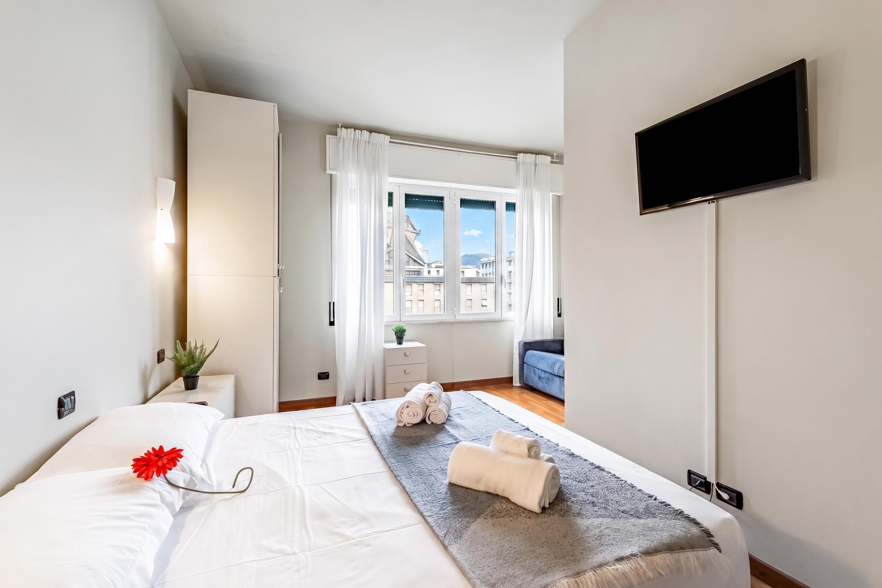 Une chambre d’hôtel avec un lit, une télévision, une fenêtre avec des rideaux, une petite table avec une plante et une vue sur les bâtiments environnants.