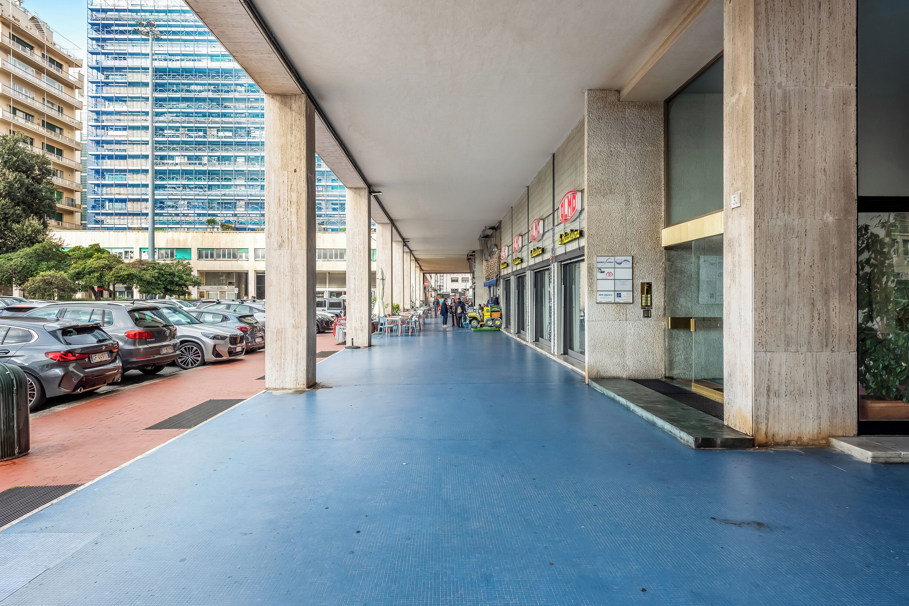 Un pasillo cubierto con coches aparcados, una hilera de tiendas y un edificio alto en construcción al fondo.