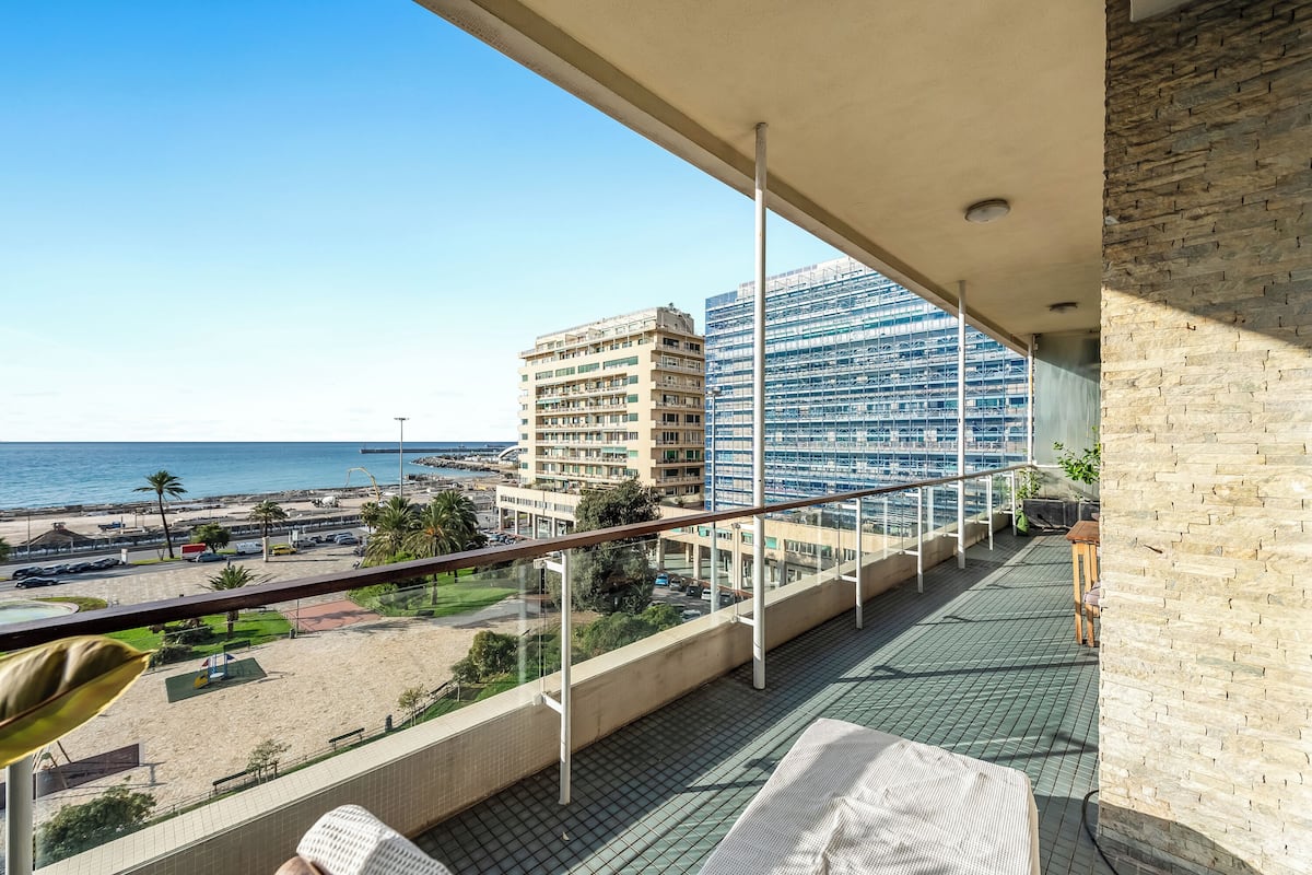 A balcony with a view of the beach and ocean, a brick wall, and a lounge area with a table and chairs.