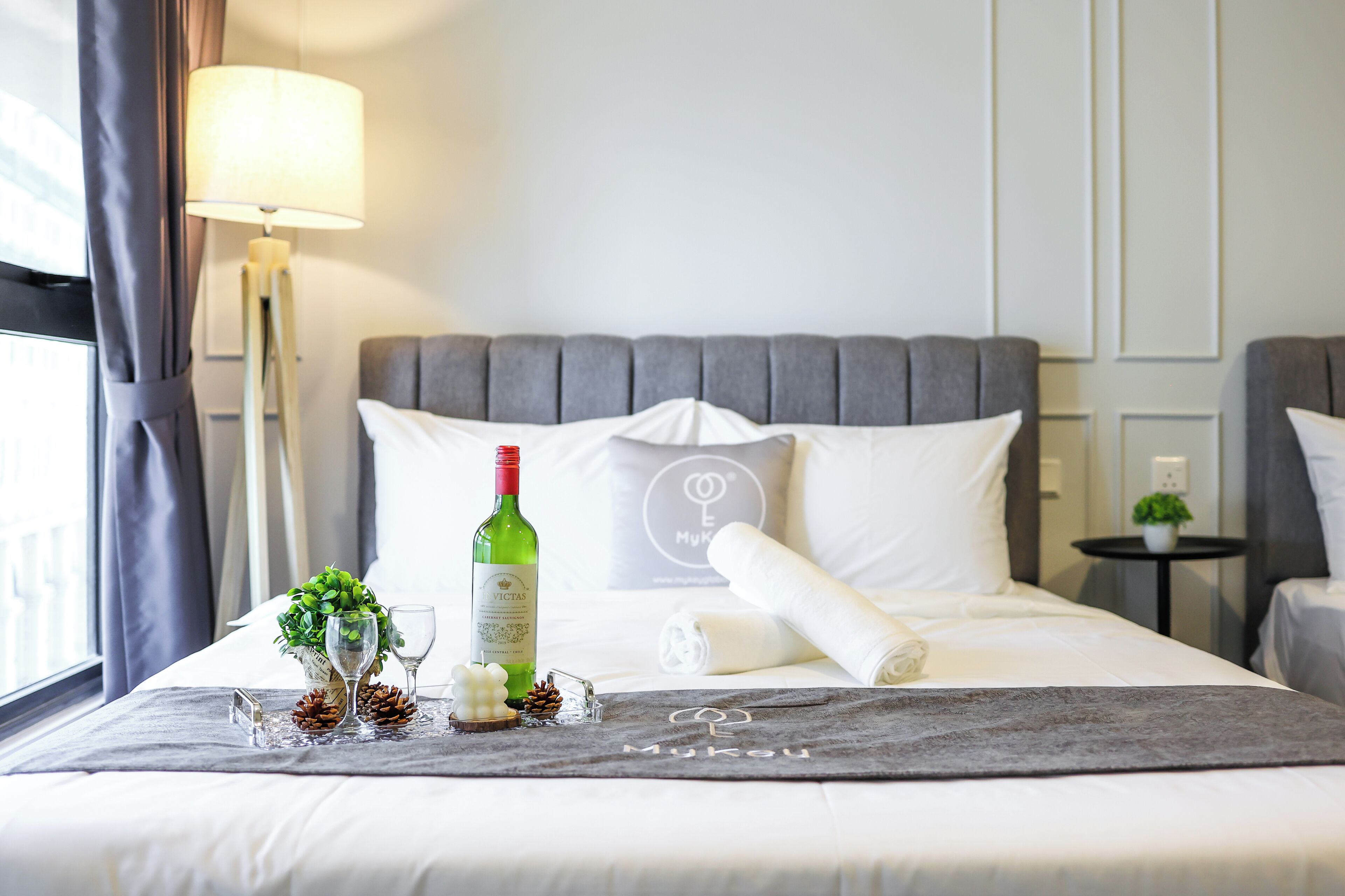 A neatly made bed with a bottle of wine, glasses, pinecones, and a towel on a bed runner with the name "Mukau".