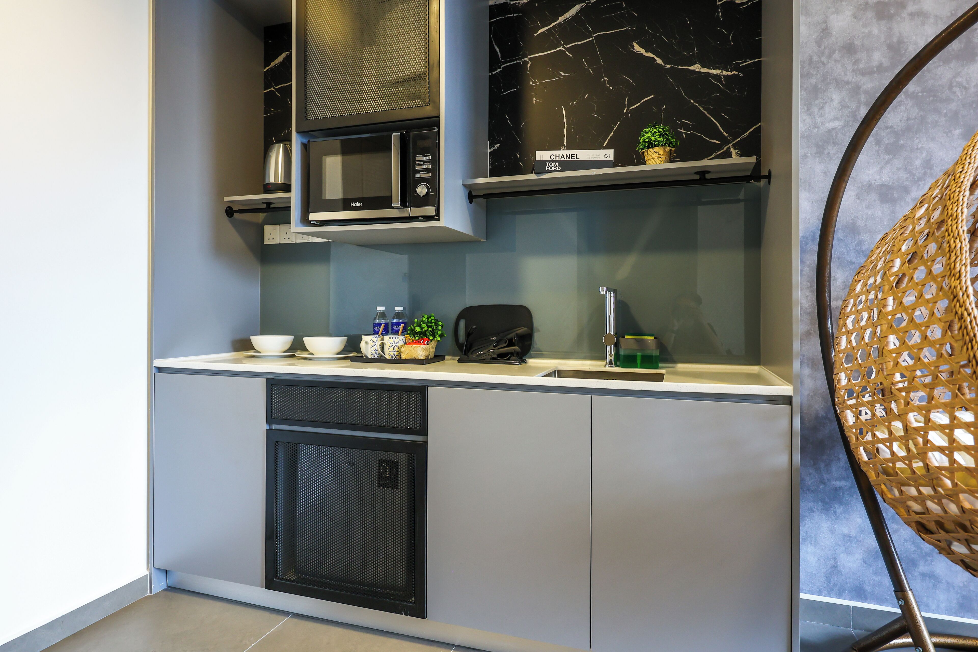A compact kitchenette with a microwave, small refrigerator, and a sink. There is a wicker chair and a potted plant on the counter.