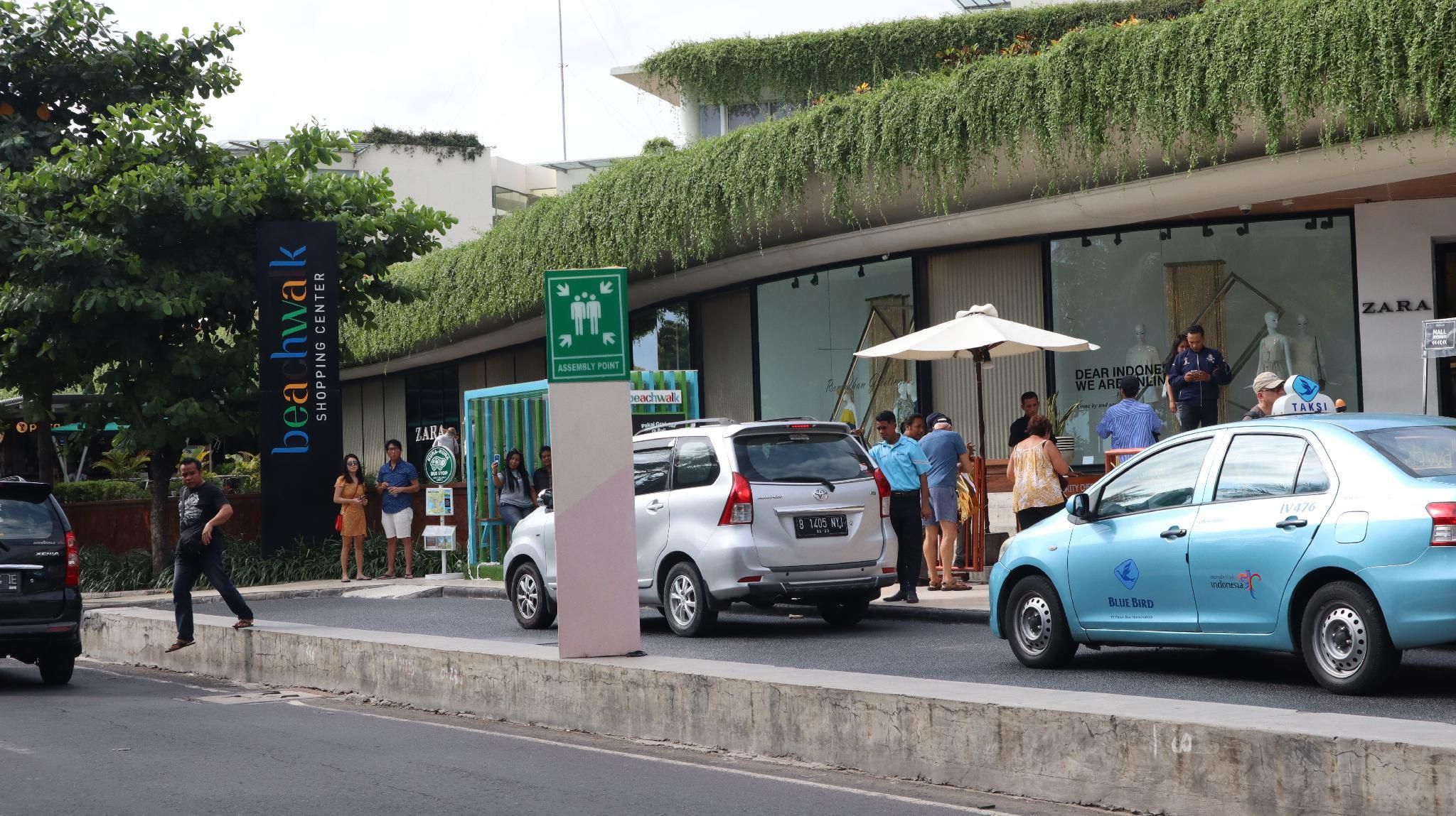 A shopping center with a green sign indicating an assembly point, a Zara store, and a Blue Bird taxi.