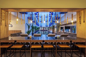 Un espacio interior amplio con una mesa larga y numerosas sillas, una escalera con barandillas azules y lámparas de madera colgantes.