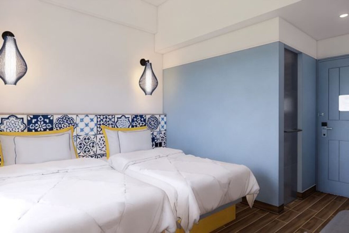 Habitación de hotel con cama, dos lámparas fijadas a la pared, cabecera estampada y puerta azul.