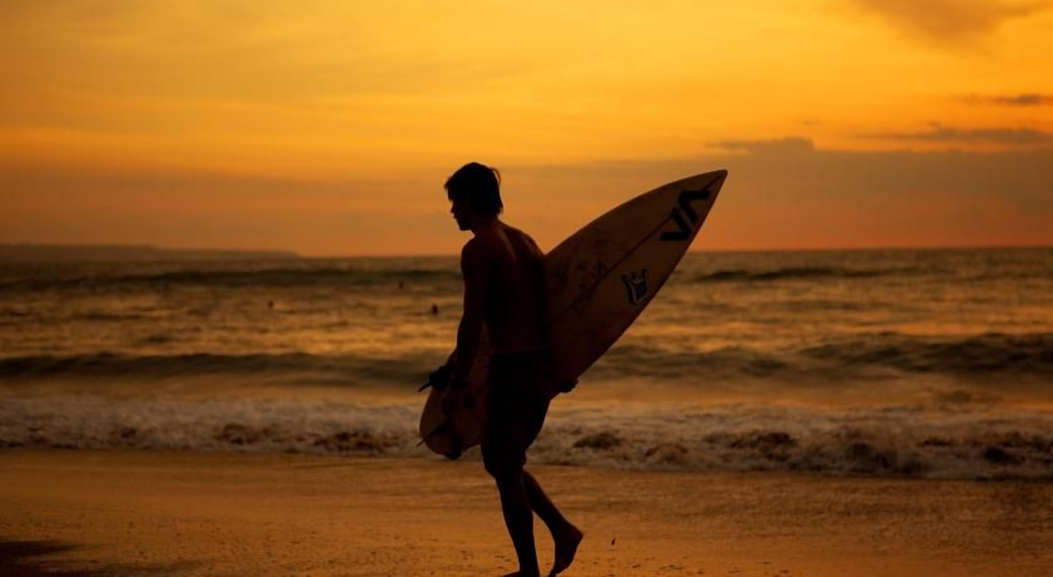 A silhouette of a person carrying a surfboard walking on a beach at sunset.