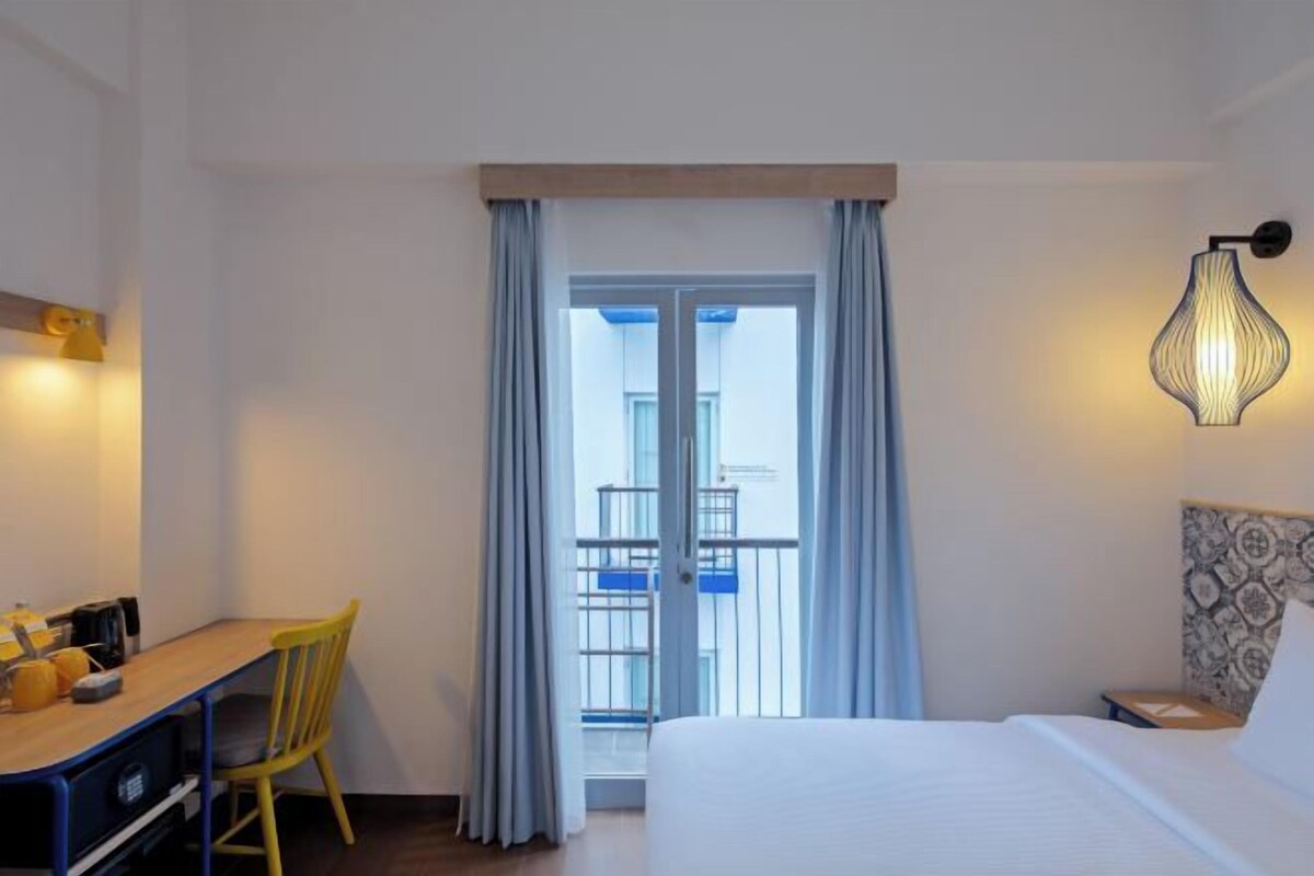A hotel room with a bed, a desk with a chair, a balcony with curtains, and a wall-mounted lamp.