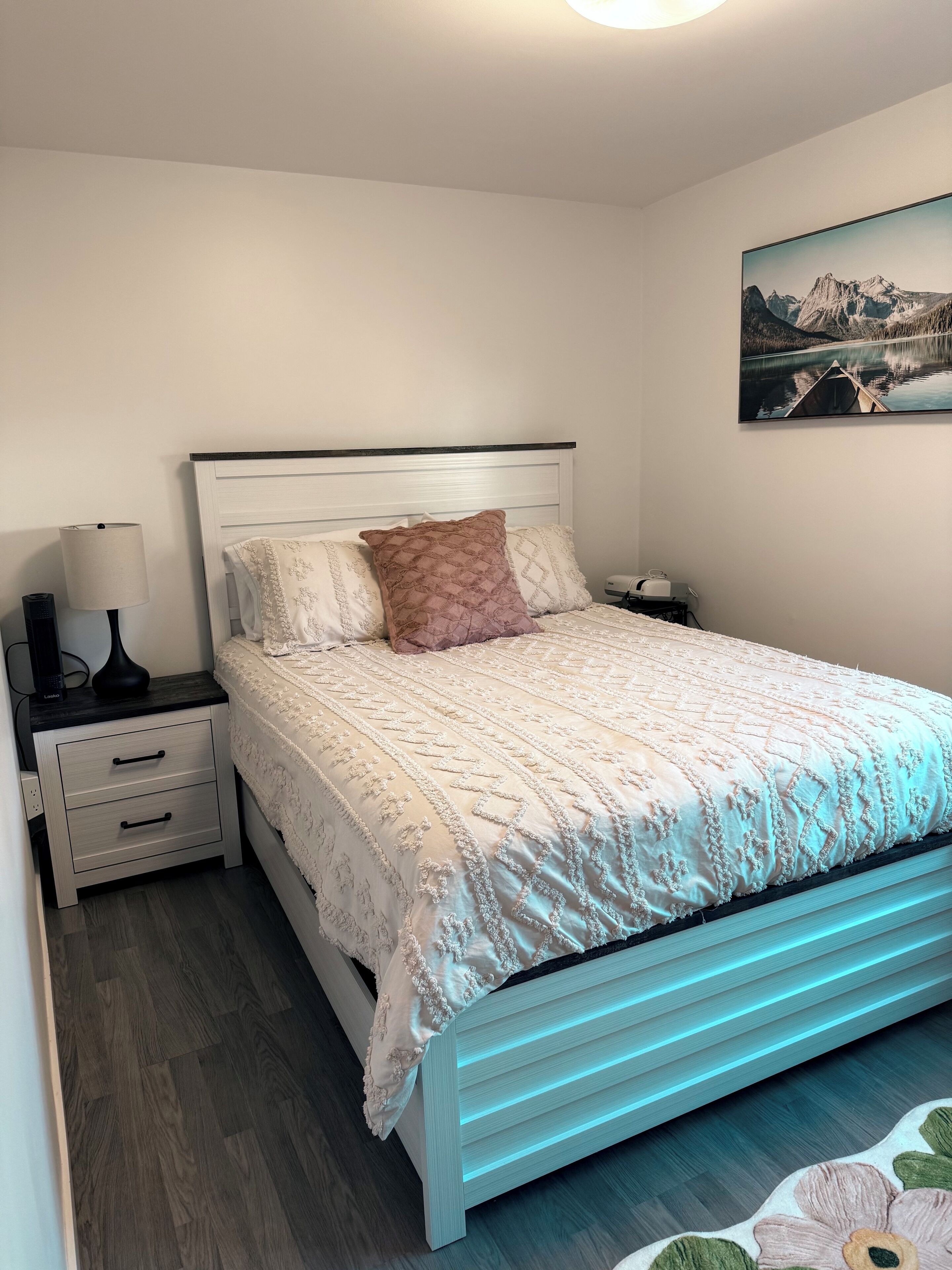 Un dormitorio ordenado con cama, mesita de noche, lámpara y un cuadro en la pared.