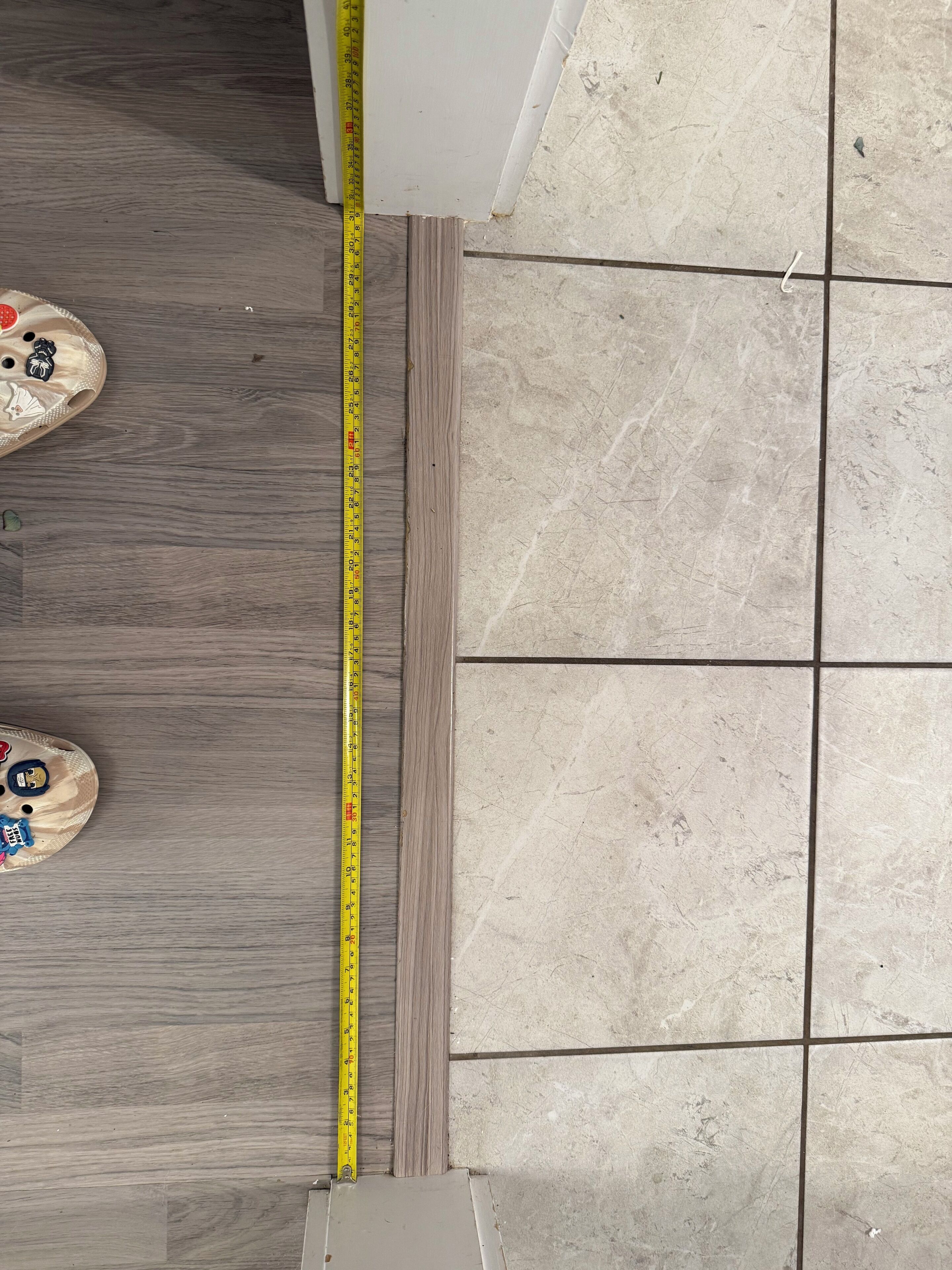 A measuring tape against a tiled floor and wooden floor, indicating room dimensions.