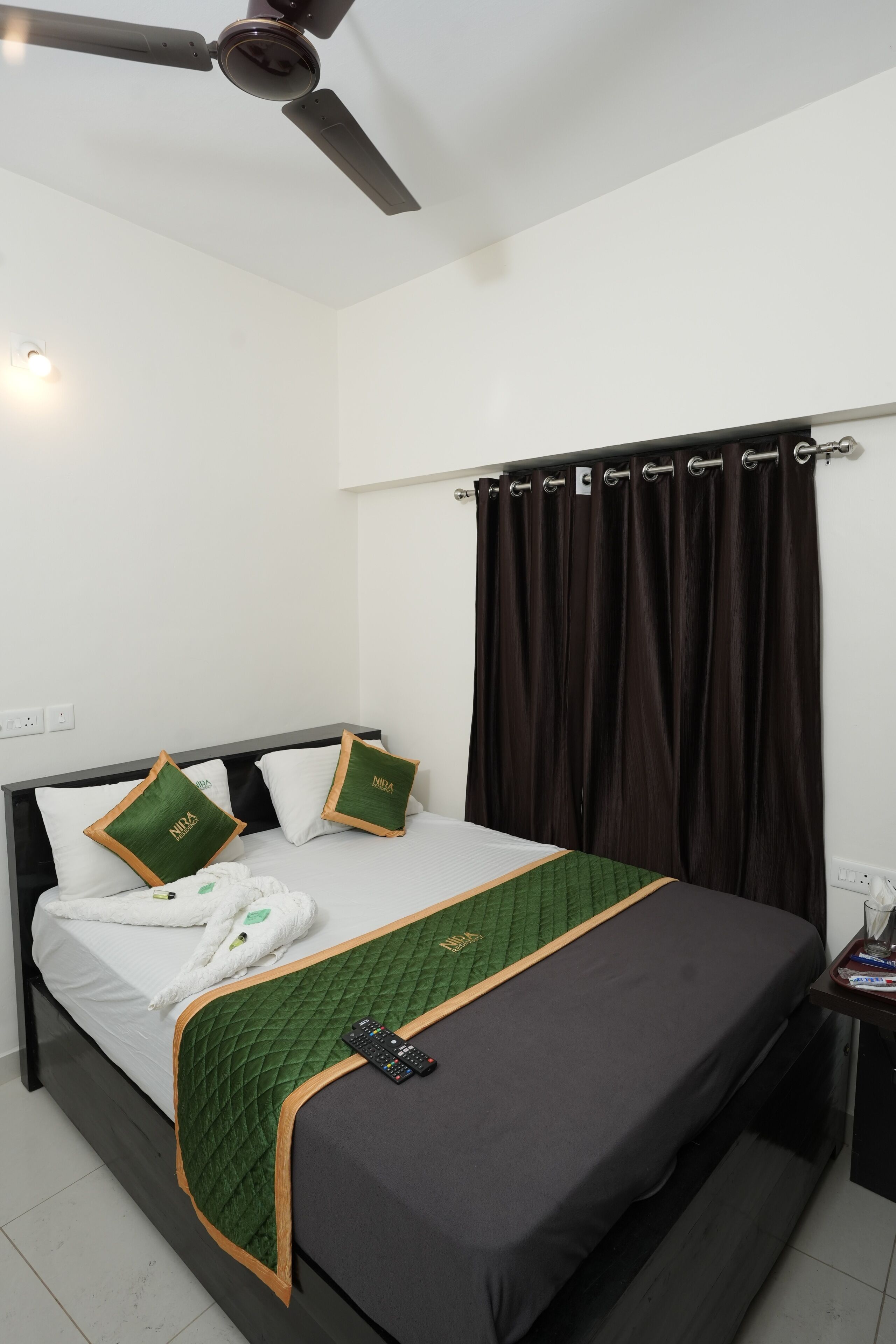 A neatly made bed with a green quilt, a remote control, and a pair of slippers on the bed.