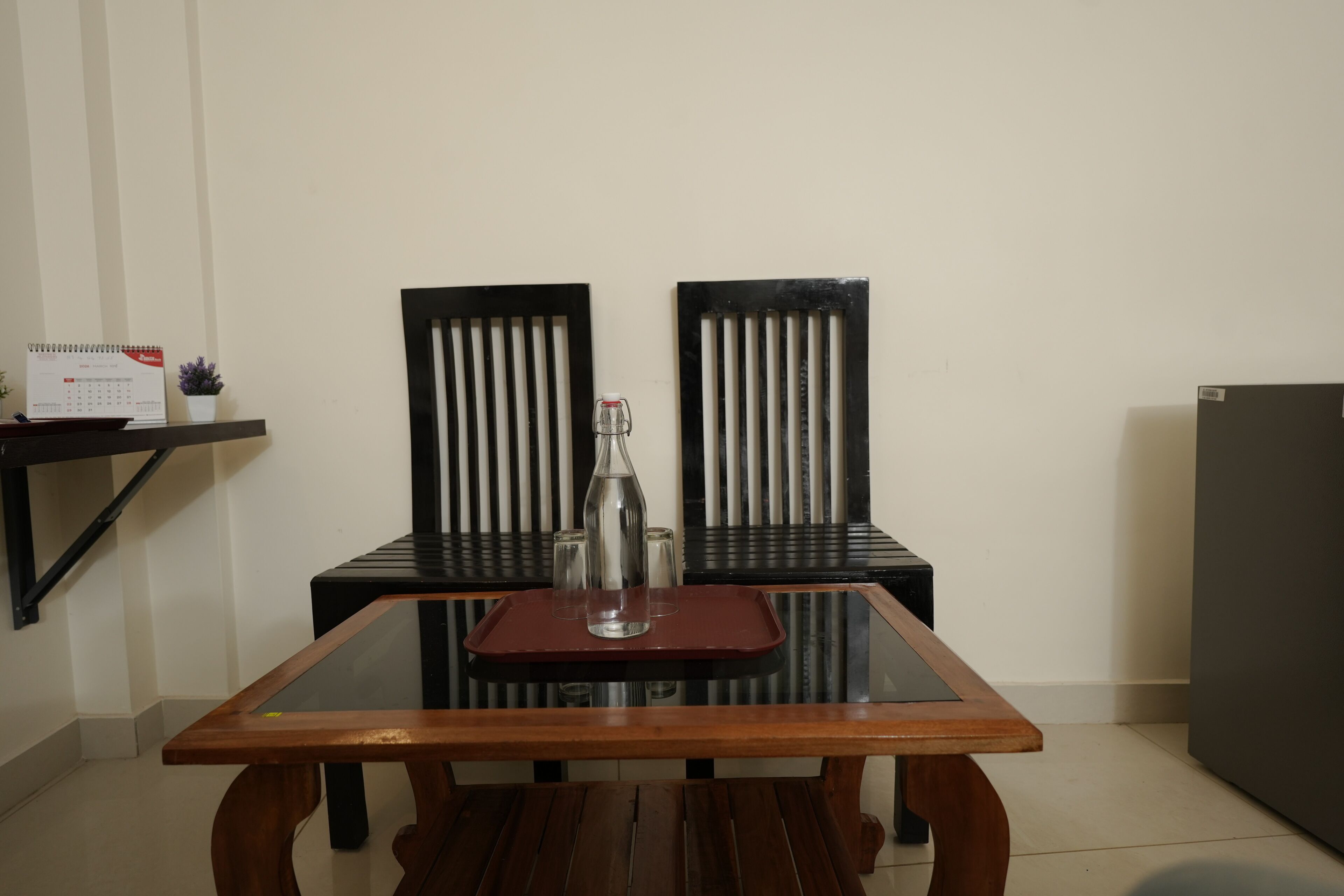 Una mesa de comedor con sillas, un calendario y una pequeña planta en maceta sobre una estantería.