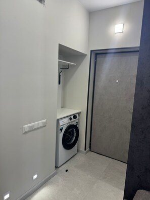 A compact laundry room with a front-loading washing machine, a small shelf, and a door. - Cozy 1-bedroom apartment in serene Yerevan with AC (Yerevan)