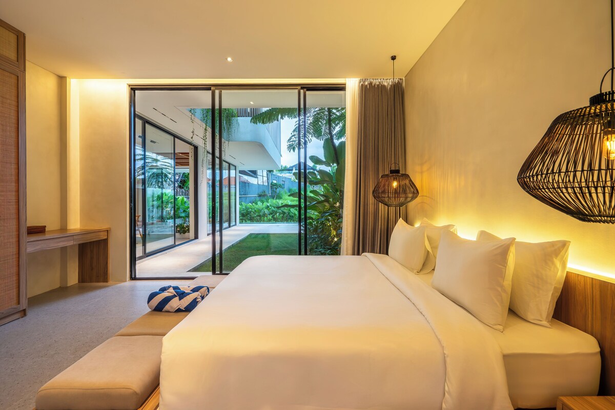 Una habitación de hotel moderna con una cama grande, un banco y vistas a un jardín exuberante a través de puertas de cristal.