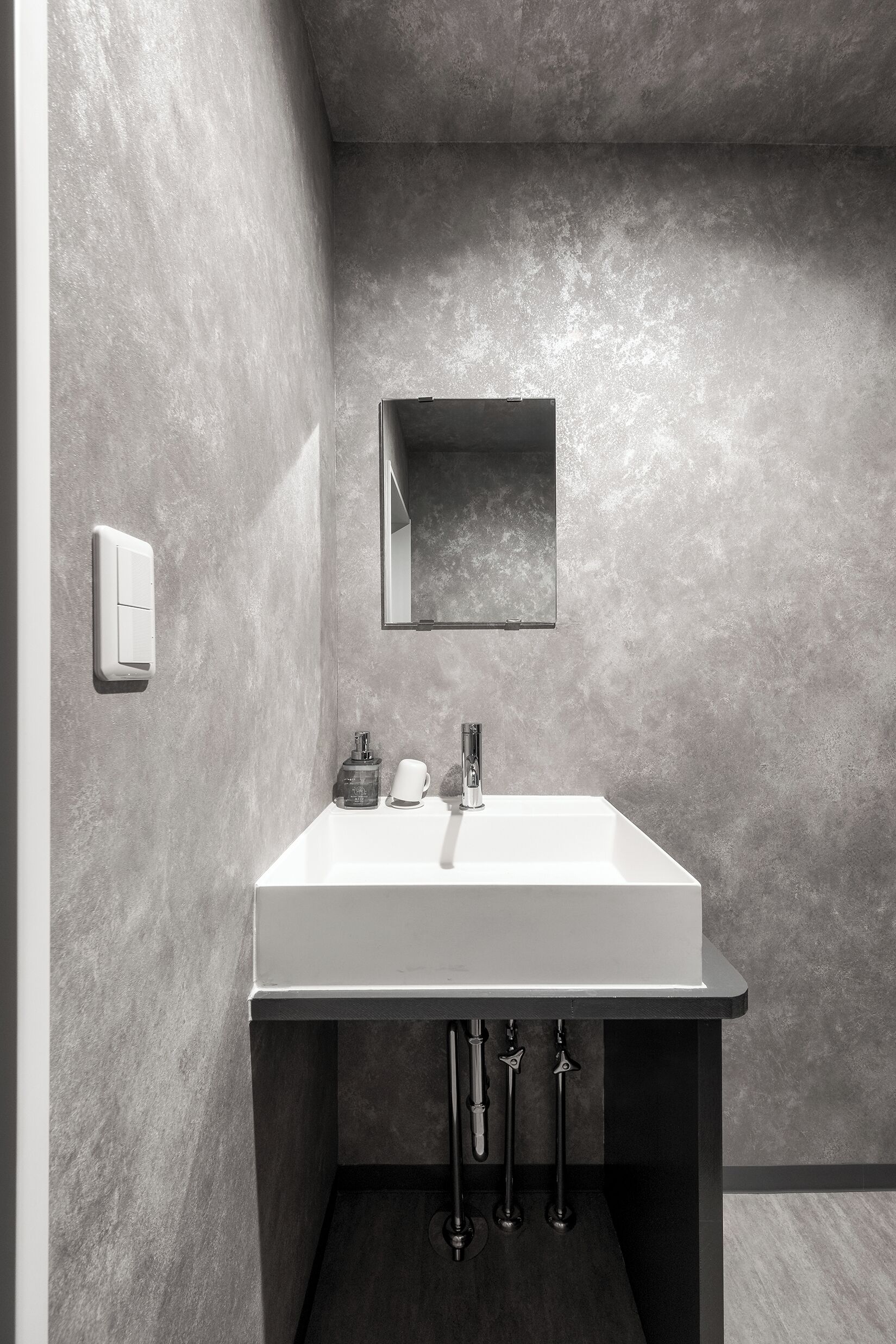 A modern bathroom with a rectangular sink, a small mirror, and a soap dispenser on the wall.