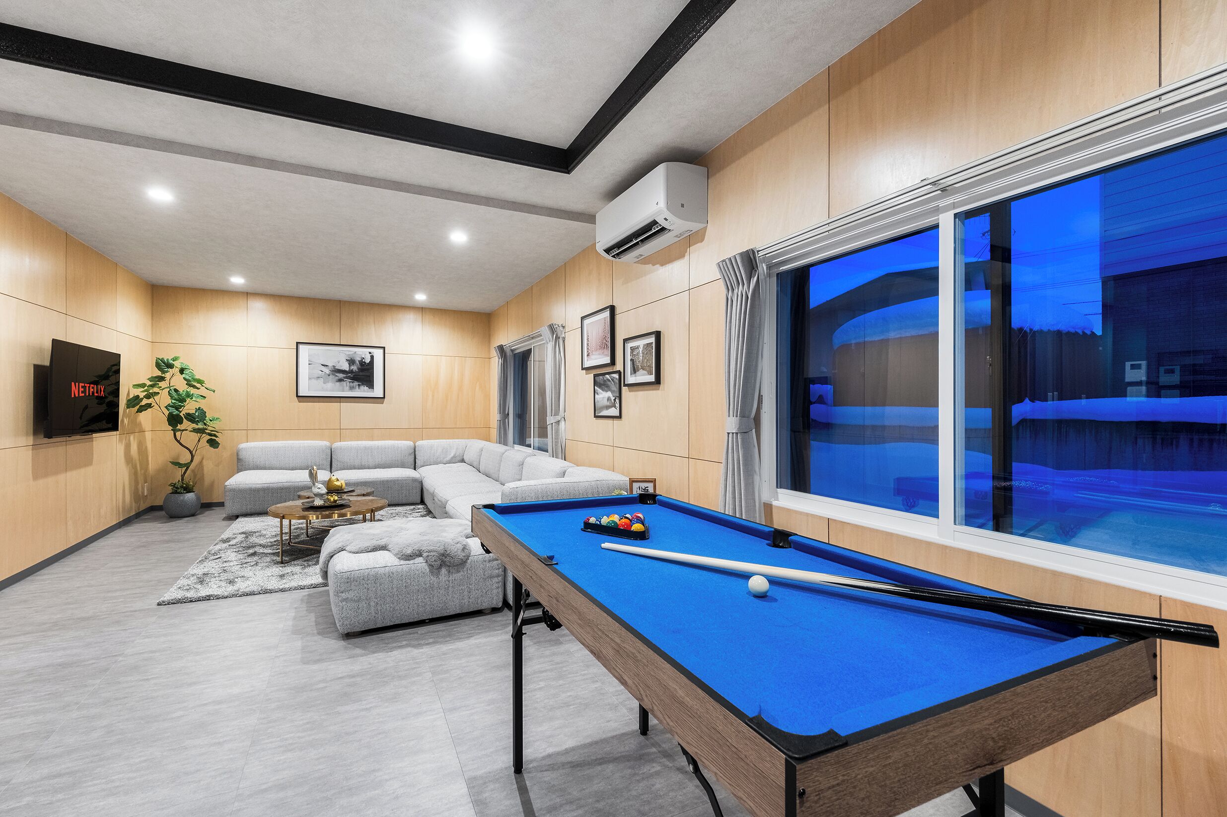 Una sala de estar moderna con mesa de billar, un sofá modular gris y un amplio ventanal con cortinas.