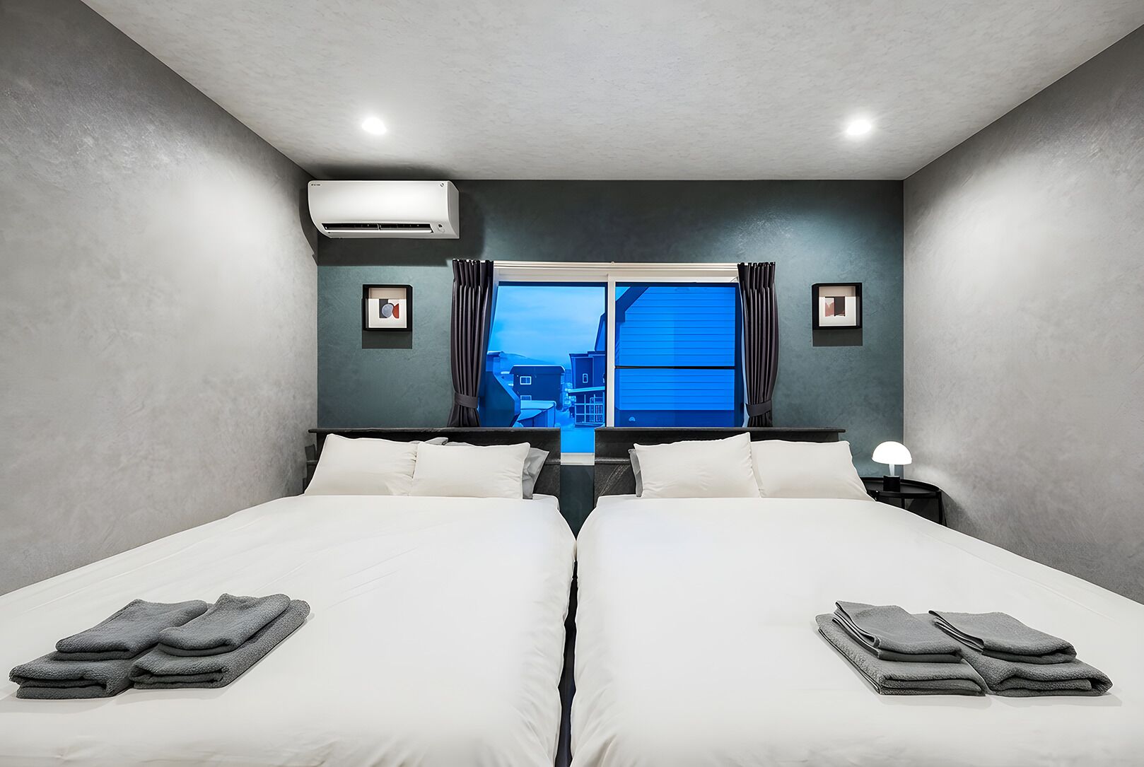 A hotel room with two beds, a window with curtains, an air conditioning unit, and a nightstand with a lamp.