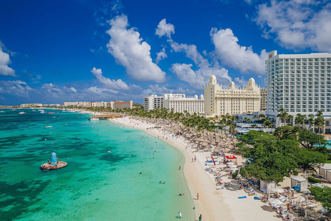 Elevated view of a bustling tropical beach resort lined with white high-rise hotels, palm trees, and turquoise water.