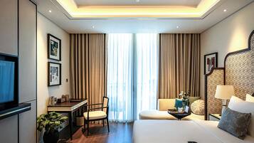 A hotel room with a bed, a desk, a chair, a TV, a sofa, and a window with curtains.