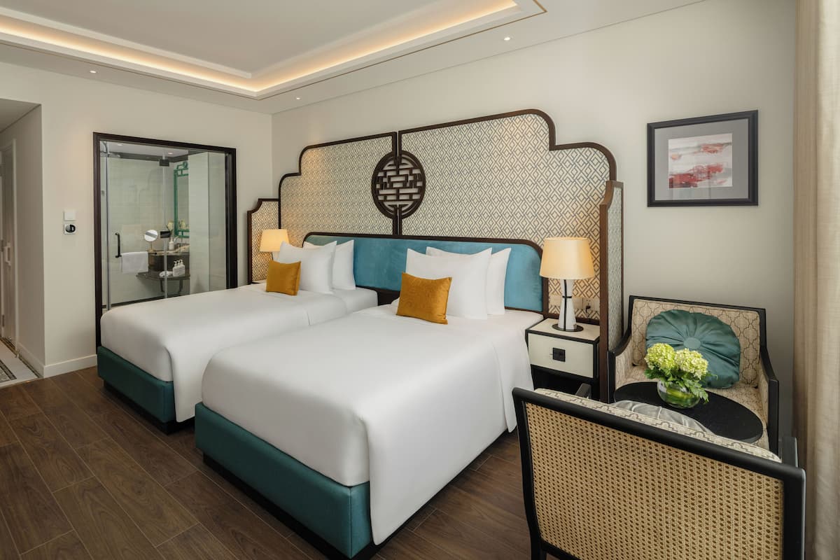 Ein Hotelzimmer mit zwei Betten, einer Sitzecke mit einem Sessel, einem Nachttisch, einer Lampe, einem gerahmten Bild an der Wand und einer Duschkabine aus Glas.