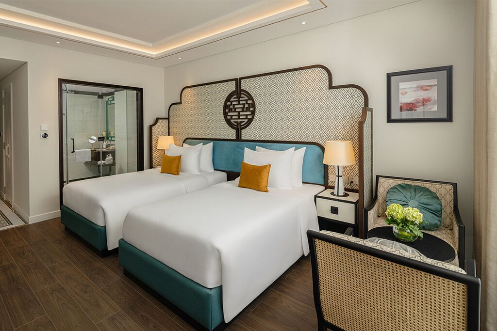 A hotel room with two beds, a glass shower enclosure, a decorative headboard, a bedside table with a lamp, and a chair with a cushion.