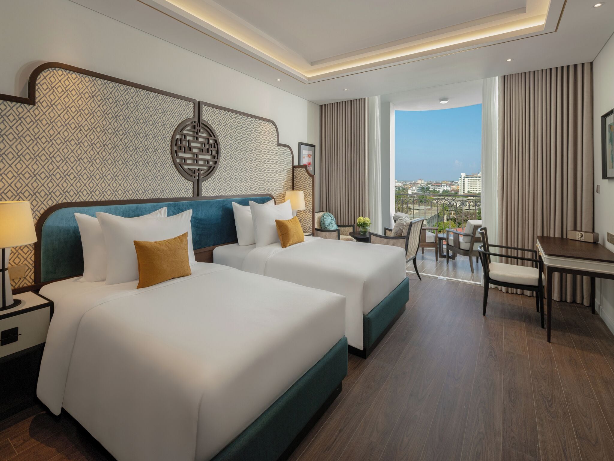 Habitación de hotel con dos camas, un escritorio y un balcón con vista a la ciudad.